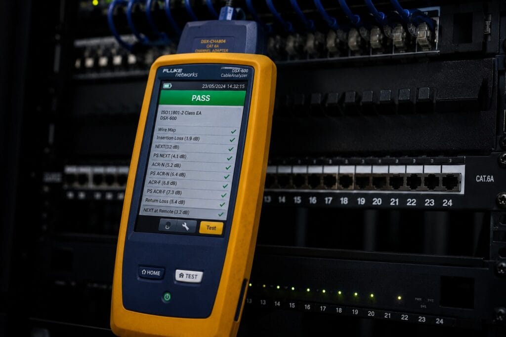 Cable analyser showing a green PASS result beside a patch panel in a commercial comms room.