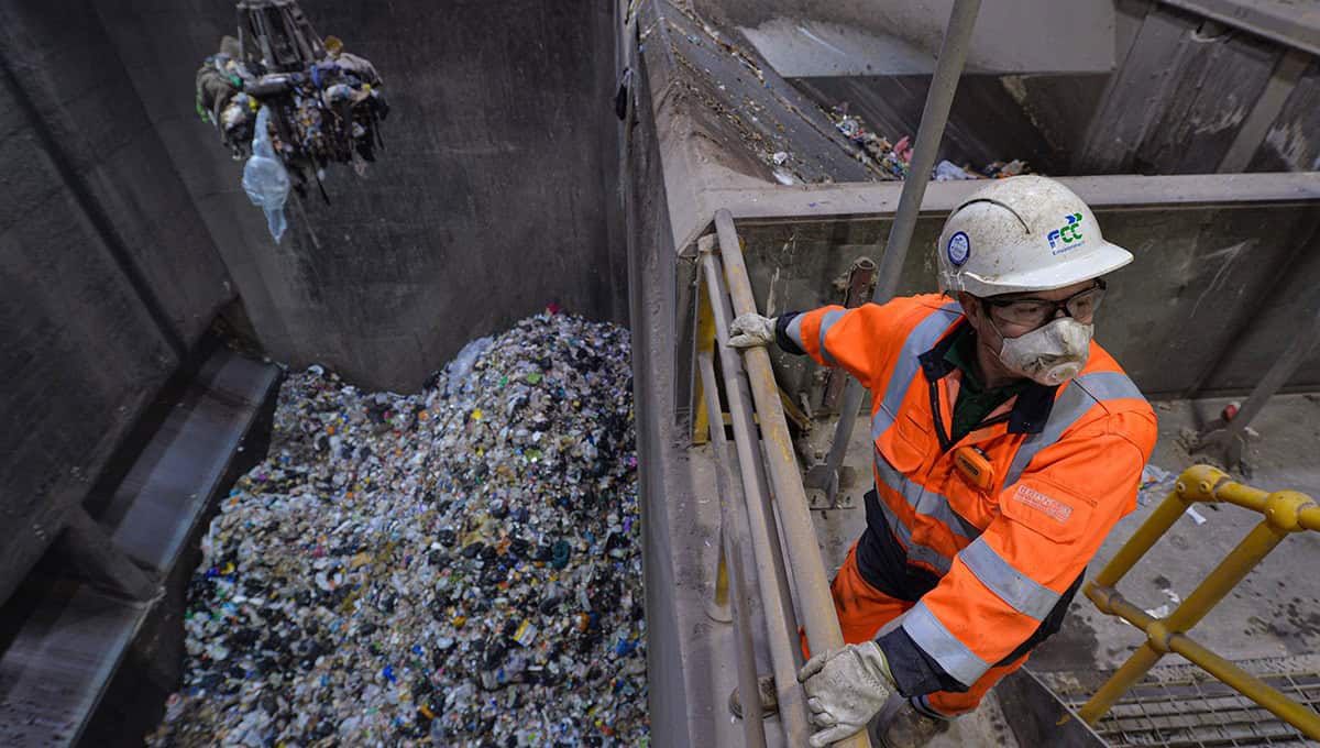 Overhead view into a waste storage silo at Kent Enviropower's Allington energy-from-waste plant, a grab crane lowering waste as a worker in an FCC-branded hard hat and dust mask looks on from a platform above the pit